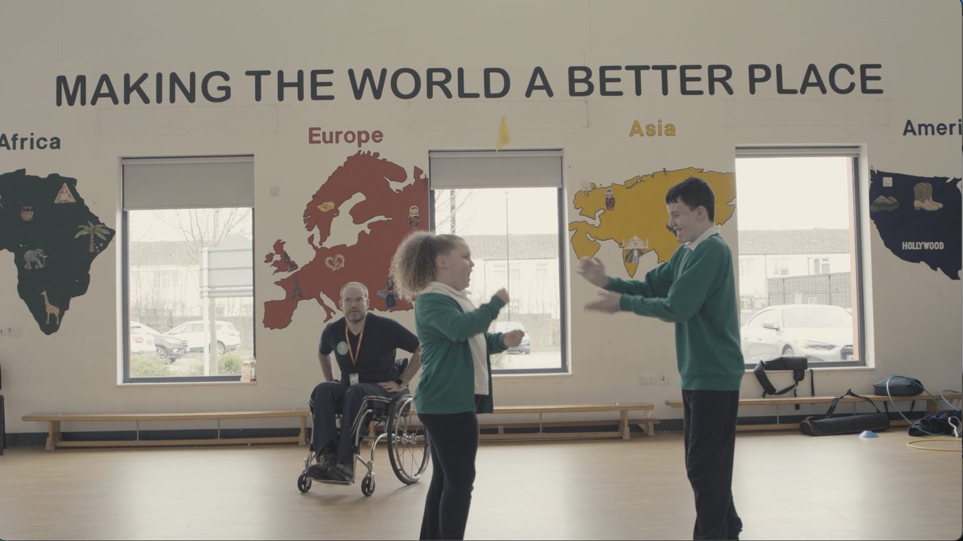 Mat Richards leading a movement session with two pupils in a school hall, beneath a wall reading Making the World a Better Place